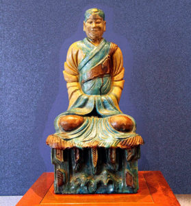 Image d'une scupture de méditant chinois, de couleur bois clair et jade sur un fond bleu