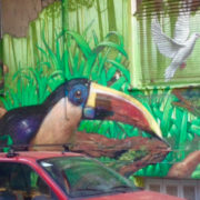 Photo de l'avant d'une voiture rouge sur fond d'un mur où est peint un toucan, de la végétation tropicale et une colombe s'envolant