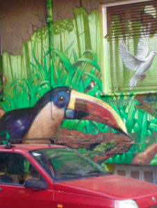 Photo de l'avant d'une voiture rouge sur fond d'un mur où est peint un toucan, de la végétation tropicale et une colombe s'envolant
