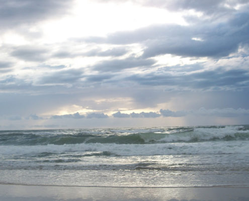 Photo d'une plage de sable que viennent battre des vagues sous un ciel nuageux percé par les rayons du soleil