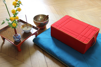 Photo d'un coussin de méditation rouge sur un tapis turquoise, avec à gauche, un petit gong et une petite table sur laquelle sont posés des fleurs en vase et un bol à encens