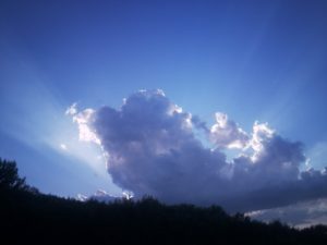 Photo d'un grand nuage, dans un ciel à diverses nuances de bleu, des bords duquel jaillissent des rayons de lumière