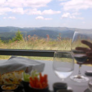 Photo d'une table de restaurant, avec une main tenant un verre à pied en premier plan, une belle vue sur une plaine fleurie et des montagnes au loin constituant l'arrière plan