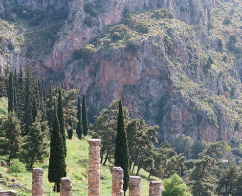 Superbe photo avec au premier plan les colonnes et vestiges en pierres d'un ancien édifice, dans un cadre naturel de collines boisées et de roches rouges