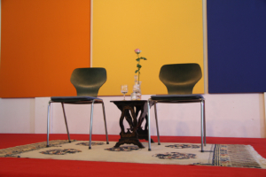 Photographie de deux chaises sur un tapis séparées par une table sur laquelle repose un soliflore avec une rose.