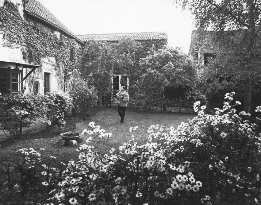 Photographie en noir et blanc où l'on voir Barbara marcher devant une maison de campagne au milieu d'un jardin en fleurs.