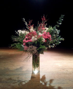 Photographie d'un bouquet de fleurs roses et blanches, dans un vase posé sur un parquet devant un fond noir et sous une lumière artificielle.