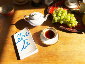 Photographie montrant, disposés sur une table, une théière, une tasse de thé, une grappe de raisins sur un plateau et le livre "Être là".