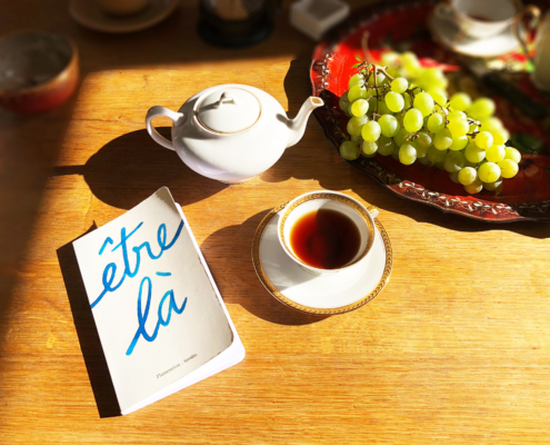 Photographie montrant, disposés sur une table, une théière, une tasse de thé, une grappe de raisins sur un plateau et le livre "Être là".