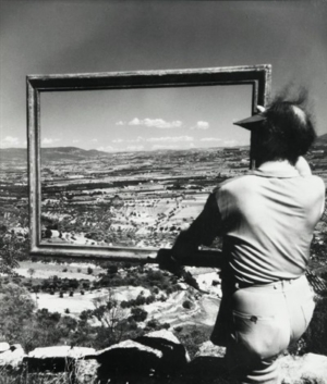 Photographie par Willy Ronis du peintre André Lhote vue de dos portant un cadre vide devant un paysage à Gordes.