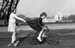 Photo noir et blanc de Robert Doiseau : un enfant en rolleur est tiré par un enfant à vélo en s'accrochant à sa jupe